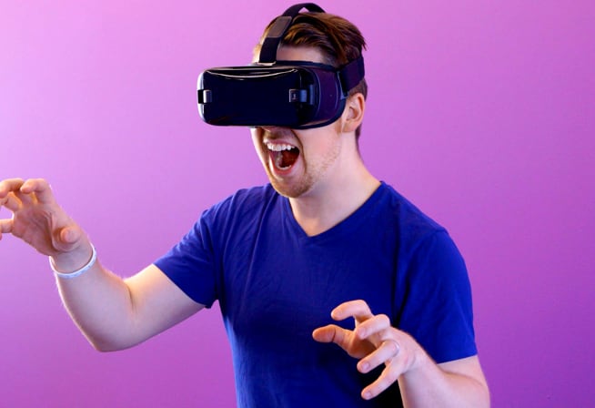 An image of white male wearing blue shirt using VR headset laughing excitedly with both arm trying to grab something